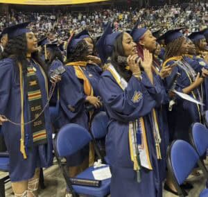 A group of graduates in blue caps and gowns stand and cheer at a graduation ceremony, with sashes and stoles visible; the venue is filled with people and a congratulatory banner hangs above.