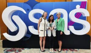 Three women stand smiling in front of a large SAS logo display. The woman in the center holds a glass award. They are dressed in professional attire, and the background features blue and white colors with modern design elements.