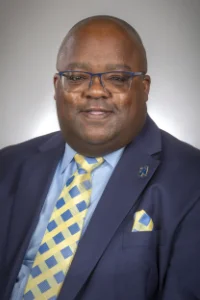 A man wearing glasses, a navy suit, a light blue shirt, and a yellow and blue patterned tie smiles at the camera against a neutral background.