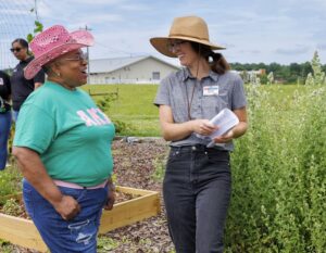 Two women stand and smile at each other in a garden. One wears a pink cowboy hat and green shirt; the other wears a wide-brimmed hat, glasses, and holds papers. Raised beds and plants are visible around them.