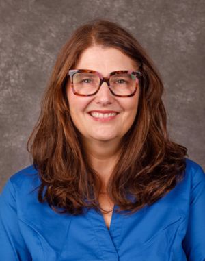 A woman with long brown hair and large, tortoiseshell glasses smiles at the camera. She is wearing a bright blue blouse and is posed in front of a gray mottled background.