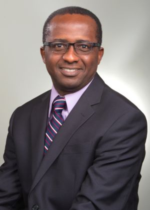 A man wearing glasses, a dark suit, a light purple shirt, and a striped tie, smiling and posing for a professional portrait against a plain, light background.