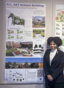 A woman in a black blazer stands smiling next to a poster displaying architectural plans and images for the N.C. A&T Science Building at North Carolina A&T State University.