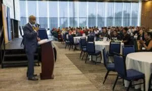 A man in a suit speaks at a podium to an audience seated at round tables in a large, bright conference room with floor-to-ceiling windows.