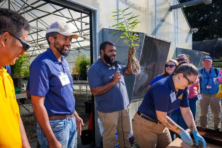 A group of people gather outside a greenhouse. One person holds up a large plant root and speaks, while others in blue shirts observe or work with plants on a table. It’s sunny and everyone looks engaged.