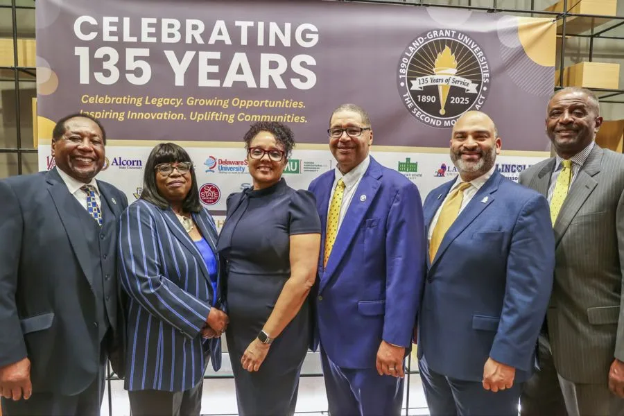 Six professionally dressed people stand together smiling in front of a banner that reads “Celebrating 135 Years” with a logo for the 1890 Land-Grant Universities and their anniversary.