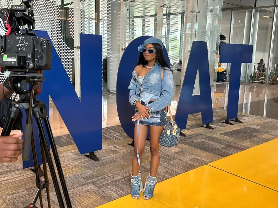 A stylish woman in denim poses for a camera in front of large blue letters spelling NCAT inside a modern building with glass walls.