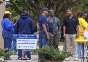 A group of people stand outside, talking and smiling near a sign for the College of Agriculture and Environmental Sciences at North Carolina A&T State University. Trees and a table with yellow bags are visible in the background.