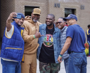 Five men smiling and posing for a selfie outdoors, wearing casual clothes including vests and caps, standing in front of a brick building.