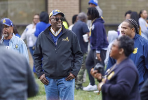 A group of people gather outdoors at an event. A man in the center wears glasses, a cap, and a black jacket, smiling as he talks with others. The background is blurred, with several people standing and chatting.