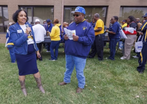 A woman speaks into a microphone while a man holding papers listens. They stand on grass in front of a brick building, surrounded by a group of people wearing yellow and blue clothing.