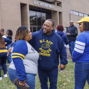 A man and woman smile and hug outside B.C. Webb Hall, surrounded by other people. The man wears a N.C. A&T Aggies Animal Science hoodie, and everyone appears cheerful at the outdoor gathering.
