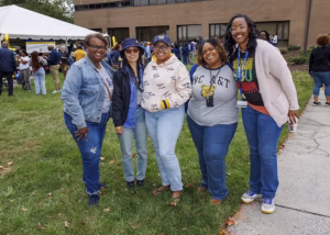 Five women smiling and posing together outdoors on grass, dressed casually in jeans and college-themed shirts and jackets, with a building and a white event tent in the background.