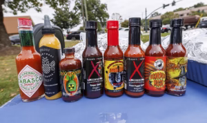 Eight bottles of various hot sauces, including Tabasco, Heartbeat Sauce, Da Bomb, and several others with colorful, fiery labels, are lined up on a blue surface outdoors with trees and cars in the background.