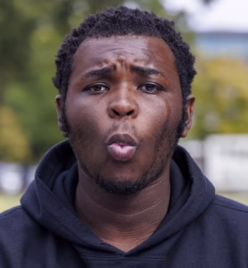 A man with short curly hair and a beard wears a black hoodie and looks directly at the camera with his lips rounded, as if whistling or surprised, outdoors with blurred greenery in the background.