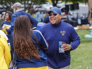 A man wearing sunglasses, a cap, and an NC A&T pullover smiles and chats with a woman outdoors at a gathering, holding a drink cup. Other people and trees are visible in the background.