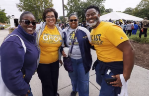 Four people stand together outdoors, smiling at the camera. They wear blue and gold clothing with GHOE and A&T logos, indicating a university event. Other people and a large white tent are visible in the background.