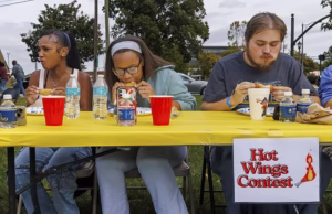 Three people sit at a table with a yellow tablecloth, eating chicken wings during a hot wings contest. Bottled water, cups, and napkins are on the table. A Hot Wings Contest sign is visible in front.