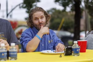A man with wavy hair, wearing a blue shirt, sits at a table outdoors and licks his finger while looking at the camera. There are water bottles, a paper plate, and a red cup on the yellow tablecloth.