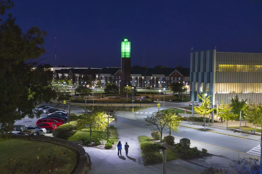 A well-lit campus scene at night featuring two people walking on a path. In the background, a tall green-lit tower stands near a modern building, with parked cars and trees in the surroundings.