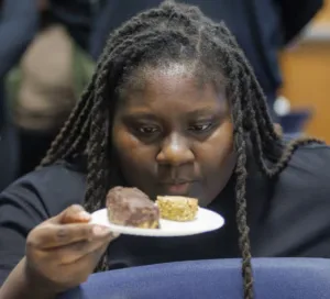 A person with long, braided hair examines two desserts on a plate, looking contemplative. The background is blurred, focusing on the individual and the treats.