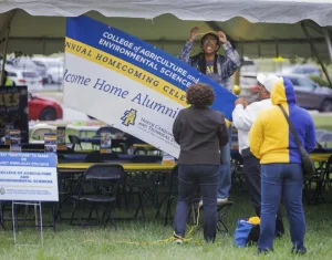 A group sets up a banner for the College of Agriculture and Environmental Sciences homecoming celebration. One person hangs the sign while others assist underneath a tent.