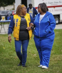 Two women walk together, smiling and engaged in conversation. One wears a yellow and blue varsity jacket, while the other is dressed in a blue tracksuit. They appear joyful in a grassy outdoor setting.