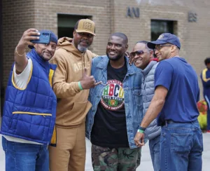 Five men pose together for a selfie outdoors, smiling and making gestures. They are dressed casually in various styles, with a brick building in the background.