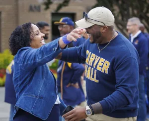Two people joyfully embrace outdoors, smiling widely. The woman wears a denim jacket, while the man sports a navy blue sweatshirt with "CARVER HALL" printed on it and a cap. Others are seen in the background.