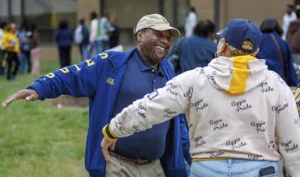 A man in a blue jacket and cap shares a joyful moment with a woman in a beige hoodie, both smiling and reaching out to each other amidst a lively outdoor gathering.
