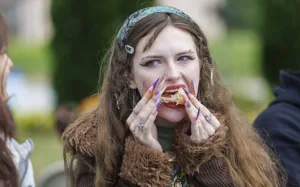 A person with long, colorful nails bites into a piece of food while looking at the camera. They have multiple piercings and wear a furry brown jacket. A blurred figure is visible in the background.