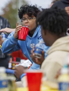A young person with curly hair and glasses sips from a red cup while seated among friends. The background features blurred figures and outdoor elements, indicating a casual social gathering.