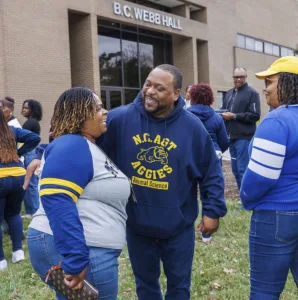 A man and woman smile at each other while engaging in conversation outside B.C. Webb Hall. Others gather in the background, creating a lively atmosphere. The woman wears a gray and blue shirt, and the man sports a blue hoodie.
