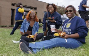 Three women sit on grass enjoying food and drinks during an outdoor event near Webb Hall. One woman is holding a plate with fried chicken and corn, while another displays a beverage. A person walks by in the background.