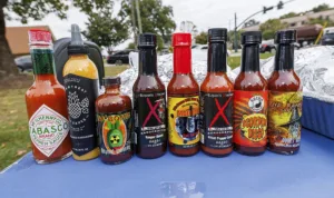 Eight bottles of hot sauce arranged on a blue tablecloth. Labels feature various flavors and heat levels, including Tabasco, Da Bomb, and Mad Dog 357, set outdoors with a blurred background of trees and vehicles.