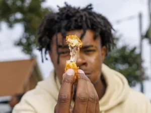 A person holds up a crispy chicken wing while looking intently at the camera. The background features greenery and hints of urban elements.