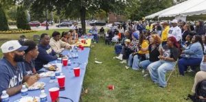 A lively outdoor gathering with people seated at long tables, enjoying food and drinks. Attendees chat and engage, with some taking photos. Various colorful outfits and beverages are present, showcasing a festive atmosphere.