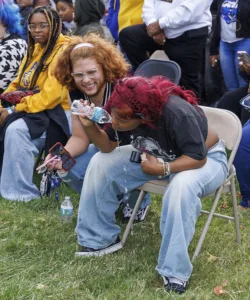 Two people sit in folding chairs at an outdoor event. One person, with curly red hair, laughs while the other, with braided red hair, leans forward, spilling water from a bottle. Others are seated in the background.