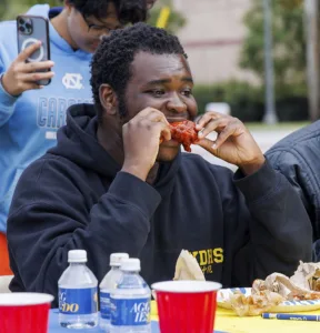 A young man is enjoying a piece of fried chicken while seated at a table covered with food debris and drinks. A person in the background is taking a photo with a smartphone.