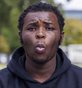A young man with short, curly hair and a slight beard makes an expressive face, with parted lips and furrowed brows, outdoors. He is wearing a black hoodie, and beads of sweat can be seen on his skin.