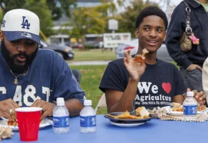 Two young men enjoy a meal at a table. One wears a blue shirt and a baseball cap, while the other, smiling, sports a black shirt with a heart and "We love Agriculture." Various food items and drinks are visible.