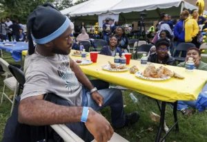A group of people enjoys a gathering, sitting at tables with food and drinks. One individual, wearing a beanie, looks pensive while others smile and engage. A festive atmosphere with tents and various attendees in the background.