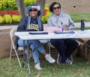 Two women sit at a table outdoors, smiling. One wears an "AGGIE" sweatshirt and a large hat; the other is in a hoodie. Various items are displayed on the table, and colorful flowers are in the background.
