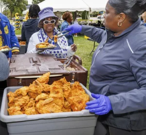 A woman in a blue hat and sunglasses receives a plate of food while another serves fried chicken from a large container. People are gathered in the background at an outdoor event.
