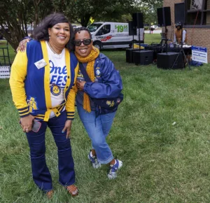Two women pose together outdoors, smiling and embracing. They wear coordinated blue and yellow outfits representing a college. In the background, there's a U-Haul truck and a DJ setup. Green grass surrounds them.
