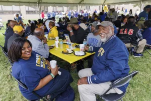 A group of people enjoying a meal under a tent, seated at tables with yellow tablecloths. Two individuals smile at the camera, surrounded by others engaged in conversation and eating.