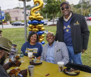 A group of people enjoying a meal at an outdoor event. Two men and a woman are seated at a table with food, while a balloon display in the background adds a festive touch.