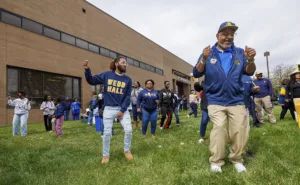A lively outdoor gathering with people dancing on the grass in front of a building. Participants wear casual clothing, including sweatshirts, and exhibit joyful expressions. The atmosphere is festive and community-oriented.