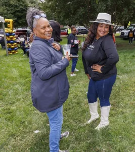 Two women smile and pose together in a grassy area during a gathering. One woman holds a drink and wears a gray jacket, while the other, in a black top and wide-brimmed hat, strikes a playful pose.