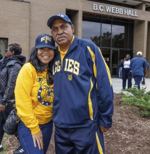 A woman and a man pose together outside B.C. Webb Hall, both wearing North Carolina A&T State University apparel. The woman smiles widely while the man has a gentle expression.